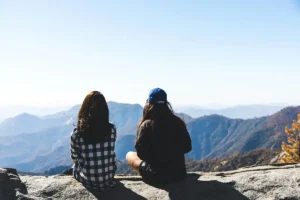 two women sit together on a rocky mountaintop, looking out over the landscape.