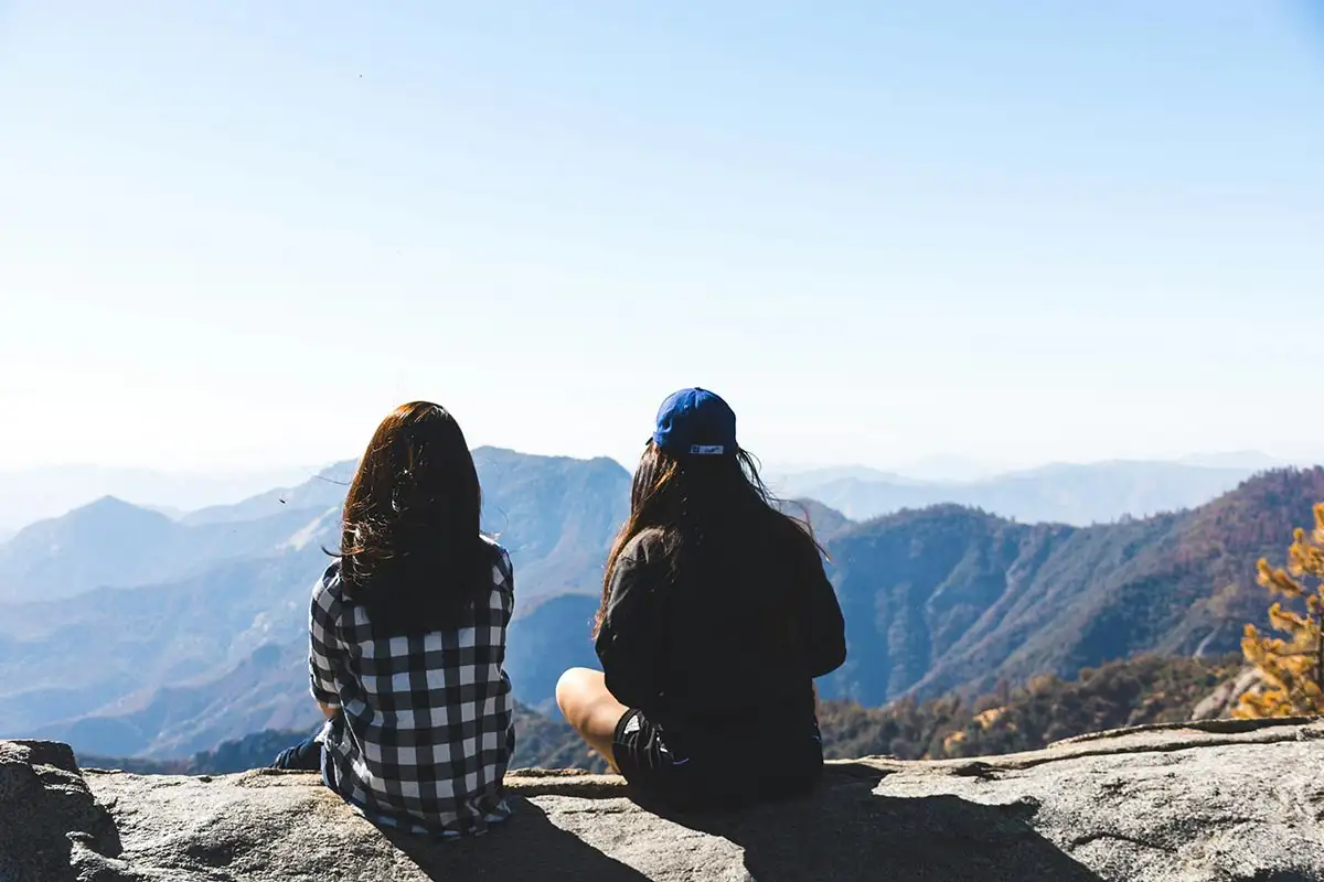 two women sit together on a rocky mountaintop, looking out over the landscape.