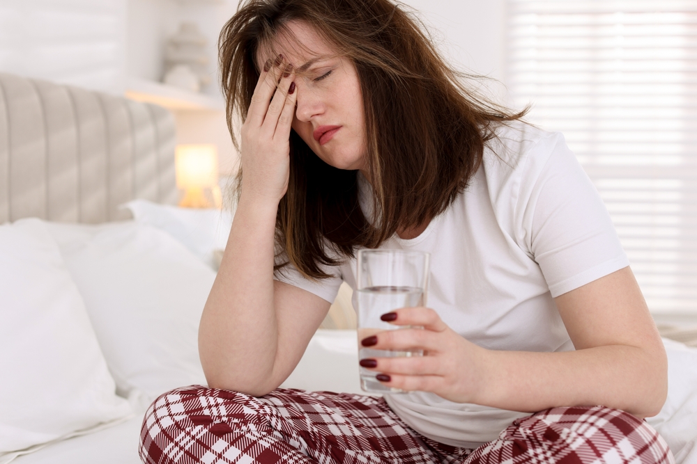 Female sitting on the bed feeling that hangover will kill her while holding a glass of water while